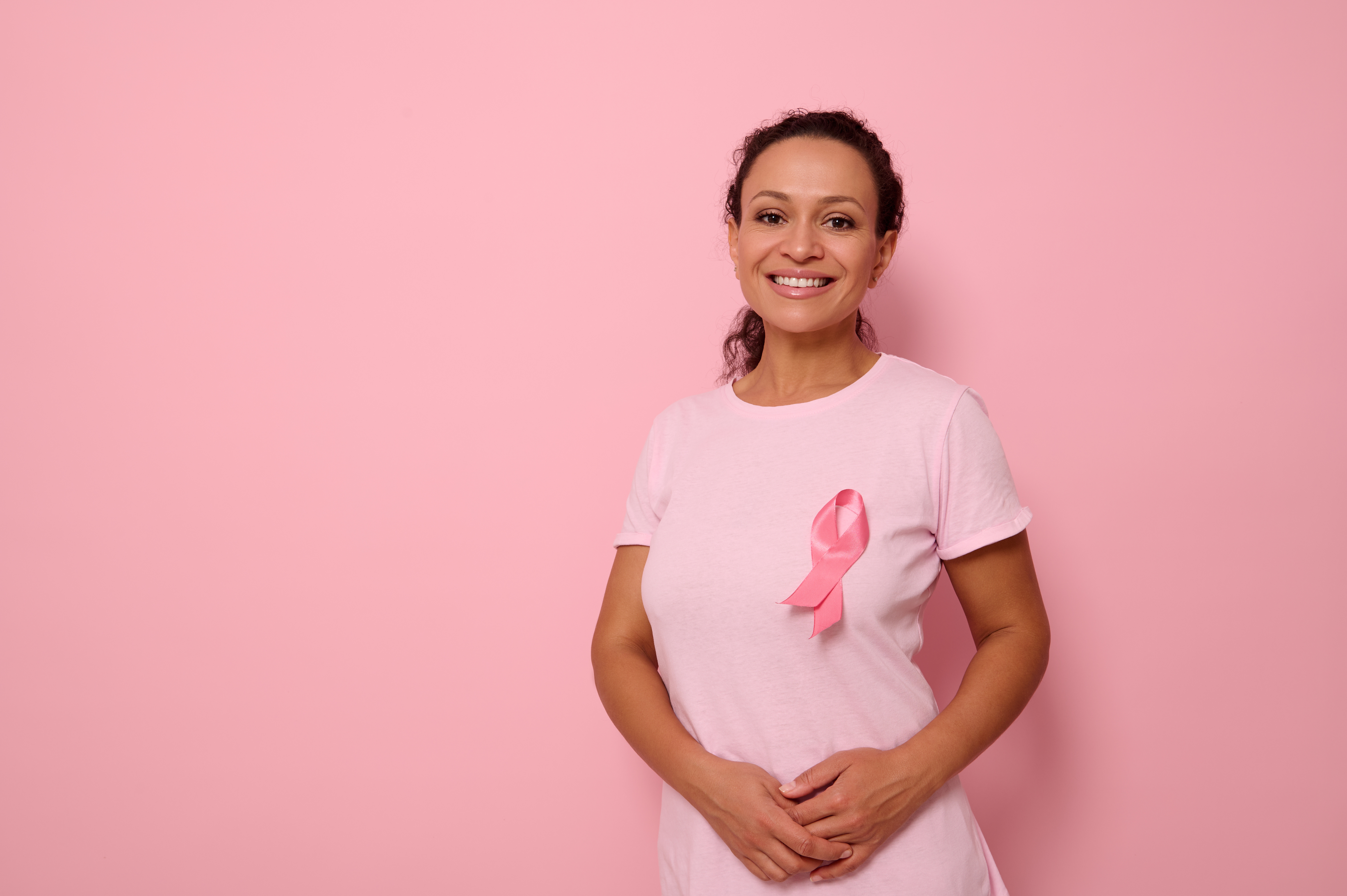Woman smiling standing up with hands crossed wearing pink t-shirt and a breast cancer ribbon on her chest